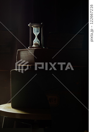 The morning sun shines hits some parts of Silver hourglass and old books in the dark room. The morning sun shines hits some parts of Silver hourglass and old books in the dark room. 122667216