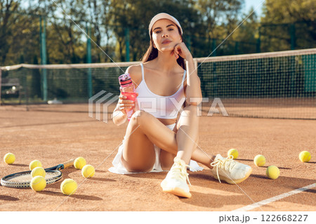 Young female playing tennis on court pausing to drink water from bottle wearing cap and sportswear 122668227