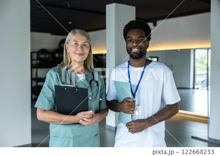 Mid aged female doctor standing with a dark-skinned intern in the clinic hall Mid aged female doctor standing with a dark-skinned intern in the clinic hall 122668233