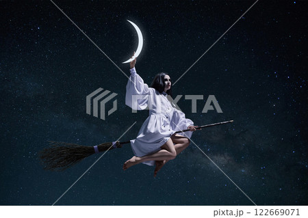 Mystical witch, woman in white gown stealing moon, flying on broomstick against dark night starry sky background 122669071