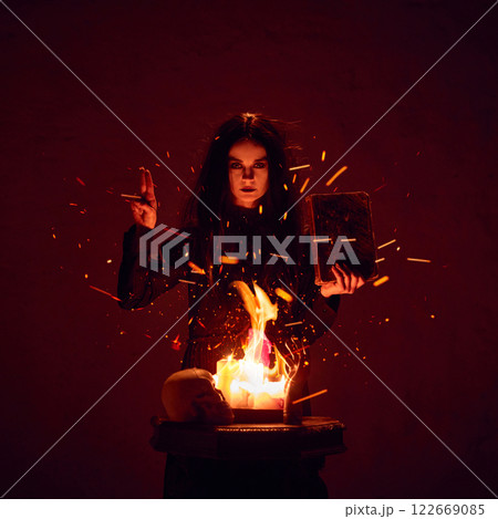 Terrifying witch, woman with dark hair, with making spells with burning fire and spell book against dark red background. 122669085