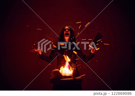 Terrifying woman with dark hair, with making spells with burning fire and spellbook against dark red background. Terrifying woman with dark hair, with making spells with burning fire and spellbook against dark red background. 122669086