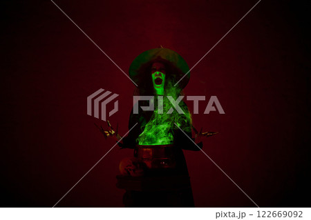 Shouting witch in black cloak and pointed hat making spell with bubbling cauldron and green mist rising around, in a dimly lit chamber. Shouting witch in black cloak and pointed hat making spell with bubbling cauldron and green mist rising around, in a dimly lit chamber. 122669092