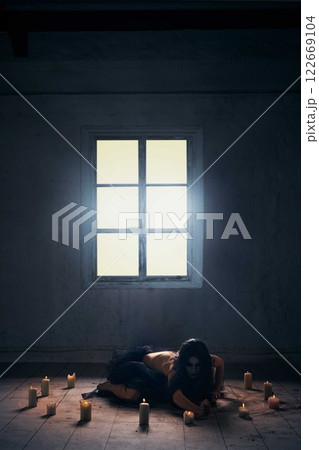 Witch ritual, spirituality, supernatural vibe. Woman covered in veil, with dark hair, sitting on floor in empty room covered with burning candles. 122669104