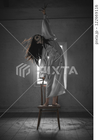 Creepy, terrifying creature, witch. Supernatural vibe. Woman with pale face, dark hair and white gown standing on chair in empty room 122669116