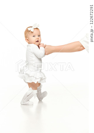 Adorable baby, girl in white outfit and cute hair accessory, holding hands with her mother and walking against white studio background 122672291