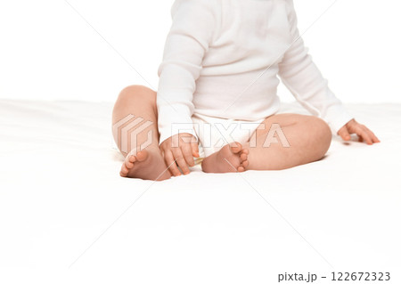 Cropped image of little girl, toddler legs and hands, baby sitting on bed in white onesie against white studio background 122672323
