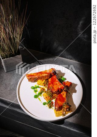 Delicious grilled pork ribs with corn and herbs served on a white plate in a dimly lit setting Delicious grilled pork ribs with corn and herbs served on a white plate in a dimly lit setting 122672692