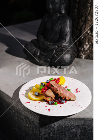 An exquisite duck dish with fresh fruit and vibrant sauce near a serene Buddha statue in an outdoor setting An exquisite duck dish with fresh fruit and vibrant sauce near a serene Buddha statue in an outdoor setting 122672807