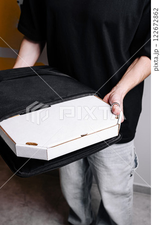 Delivery person holds pizza box in black carrying bag near a modern interior during urban evening Delivery person holds pizza box in black carrying bag near a modern interior during urban evening 122672862