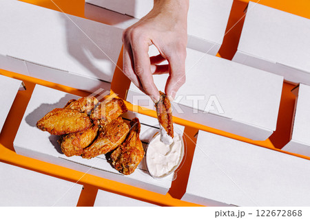 Hand dipping crispy chicken wings into creamy sauce on a bright orange surface with white boxes in the background 122672868
