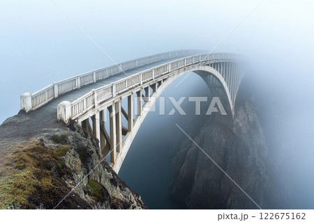 Misty wooden bridge leads to a lonely tree on a rocky island during early morning fog 122675162