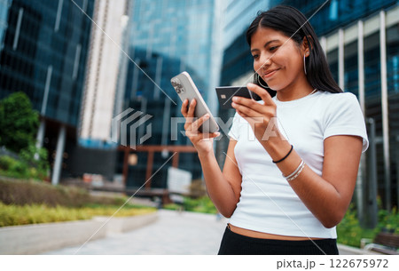 A Young Woman Actively Engaged with Her Smartphones Within a Bustling Urban Setting A Young Woman Actively Engaged with Her Smartphones Within a Bustling Urban Setting 122675972