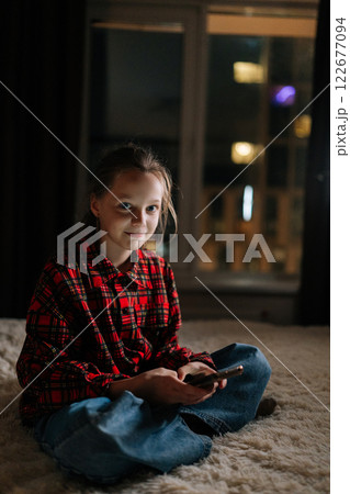 Vertical portrait of smiling young girl using smartphone at night in dark living room by window, with colorful bokeh lights on blurred background creating warm festive atmosphere, looking at camera 122677094