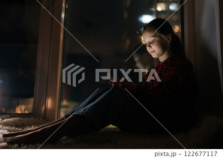 Side view of cute 10 yo child girl smiling looking to screen typing on smartphone, sitting alone at night by window colorful bokeh lighting creating vibrant nighttime ambiance on blurred background. Side view of cute 10 yo child girl smiling looking to screen typing on smartphone, sitting alone at night by window colorful bokeh lighting creating vibrant nighttime ambiance on blurred background. 122677117