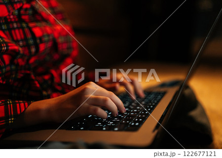 Close-up hands of unrecognizable teen girl wearing red and black checkered shirt typing on backlit laptop keyboard sitting on bed in dark room, creating cozy atmosphere. Close-up hands of unrecognizable teen girl wearing red and black checkered shirt typing on backlit laptop keyboard sitting on bed in dark room, creating cozy atmosphere. 122677121