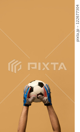 Both hands in blue goalkeeper gloves gripping soccer ball, highlighting teamwork and protection in competitive sports against beige background. Both hands in blue goalkeeper gloves gripping soccer ball, highlighting teamwork and protection in competitive sports against beige background. 122677304