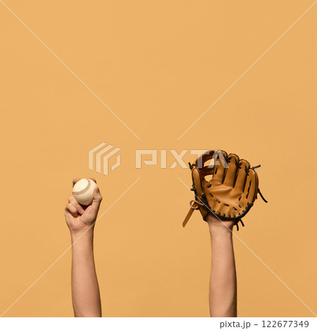Raised hand in leather baseball glove, and gripping ball, symbolizing defensive preparation and teamwork against warm-toned background. Raised hand in leather baseball glove, and gripping ball, symbolizing defensive preparation and teamwork against warm-toned background. 122677349