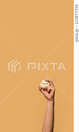 Hand holding baseball high, fingers gripping seams with precision, symbolizing control and readiness for action against warm-toned background. 122677356