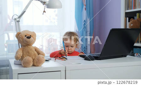 In a cozy nursery, a baby girl sits at her desk, focused on taking notes with her laptop nearby. Freelance and distance work concept In a cozy nursery, a baby girl sits at her desk, focused on taking notes with her laptop nearby. Freelance and distance work concept 122678219