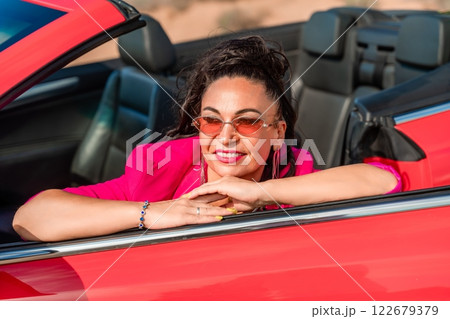 A woman in a pink jacket is sitting in a red convertible. She is wearing sunglasses and has her hand on her hip. Scene is relaxed and carefree. A woman in a pink jacket is sitting in a red convertible. She is wearing sunglasses and has her hand on her hip. Scene is relaxed and carefree. 122679379
