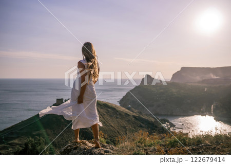 A woman in a white dress stands on a hill overlooking the ocean. The scene is serene and peaceful, with the woman's dress billowing in the wind. The combination of the ocean. A woman in a white dress stands on a hill overlooking the ocean. The scene is serene and peaceful, with the woman's dress billowing in the wind. The combination of the ocean. 122679414