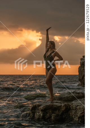 Woman Sunset Beach Pose - A woman in a black swimsuit poses on a rocky beach with the sunset in the background. Woman Sunset Beach Pose - A woman in a black swimsuit poses on a rocky beach with the sunset in the background. 122679529