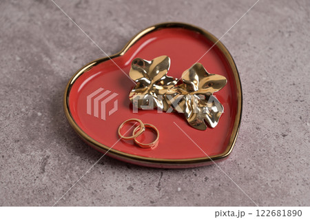 Couple of wedding golden rings and earrings on red heart shape ceramic plate with a gold rim. Gray concrete background. Valentine's Day, marriage, wedding 122681890
