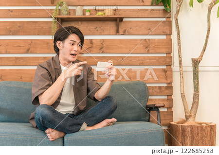 家のリビングでスマホを見ながらくつろぐ若いアジア人男性(驚く・ショック・衝撃・ビックリ) 家のリビングでスマホを見ながらくつろぐ若いアジア人男性(驚く・ショック・衝撃・ビックリ) 122685258