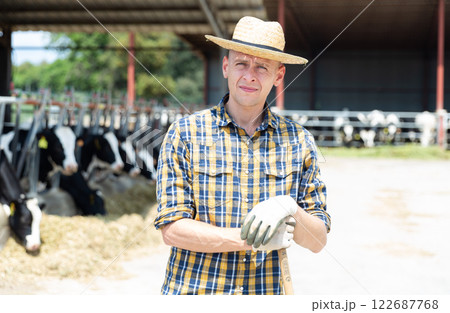 Confident owner of dairy farm standing on background with cows in stall Confident owner of dairy farm standing on background with cows in stall 122687768