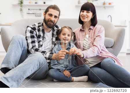 Young Caucasian family with parents and daughter celebrating new apartment. Father, mother, and child holding keys, expressing happiness and unity indoors, sitting in living room. 122692720