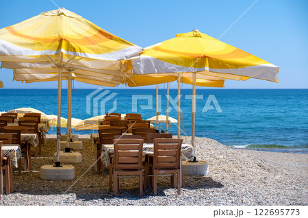 Relaxing beachside cafe scene. Wooden tables and chairs under vibrant yellow umbrellas, overlooking a stunning turquoise sea 122695773