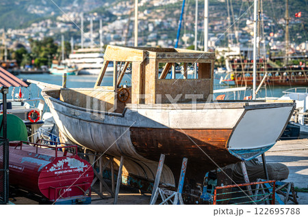 A weathered fishing boat under repair in a bustling shipyard. The hull shows signs of age and wear, contrasting with the vibrant background of other vessels and a city skyline 122695788