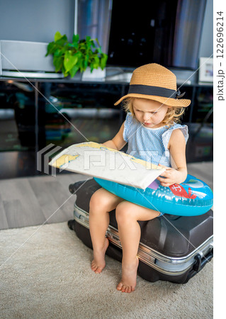 Little girl with suitcase baggage luggage and inflatable life buoy reading map and ready to go for traveling on vacation Little girl with suitcase baggage luggage and inflatable life buoy reading map and ready to go for traveling on vacation 122696214