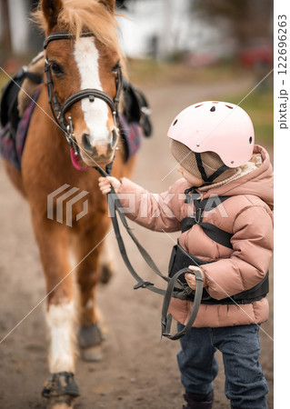 Little girl in protective jacket and helmet with her brown pony before riding Lesson Little girl in protective jacket and helmet with her brown pony before riding Lesson 122696263
