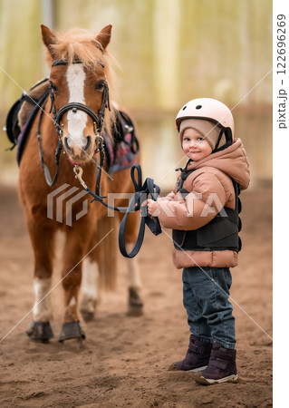 Portrait of little girl in protective jacket and helmet with her brown pony before riding Lesson Portrait of little girl in protective jacket and helmet with her brown pony before riding Lesson 122696269