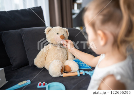 Little girl playing doctor with toys and teddy bear on the sofa in living room at home Little girl playing doctor with toys and teddy bear on the sofa in living room at home 122696296