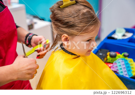 Hairdresser making a hair style to cute little girl in real child salon with colorful interior and toys Hairdresser making a hair style to cute little girl in real child salon with colorful interior and toys 122696312