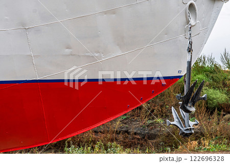 Ship's bow with a red and white hull, flanked by two large anchors on chains close-up. Vessel is docked on dry land for maintenance 122696328