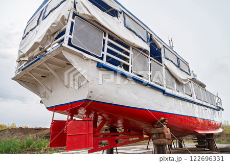 Stern of large boat on wooden supports for repair on dry dock, with covered upper deck and metal railings, low angle view against cloudy sky 122696331