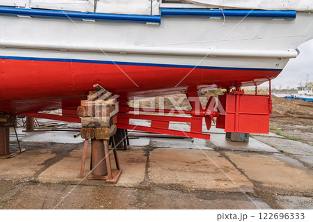 Dry-docked boat with a red and white hull, showing its rudder system and propeller, supported by wooden and metal blocks, in shipyard close view 122696333