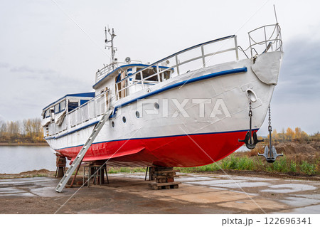 White and red ship raised on blocks for maintenance at dry dock near river, with cloudy sky and autumn trees in background. Concept of ship repair 122696341