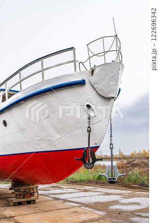 White and red ship raised on blocks for maintenance at dry dock near river, with cloudy sky and autumn trees in background. Concept of ship repair White and red ship raised on blocks for maintenance at dry dock near river, with cloudy sky and autumn trees in background. Concept of ship repair 122696342