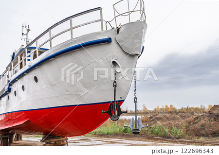 White and red ship raised on blocks for maintenance at dry dock near river, with cloudy sky and autumn trees in background. Concept of ship repair 122696343