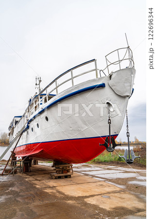 White and red ship raised on blocks for maintenance at dry dock near river, with cloudy sky and autumn trees in background. Concept of ship repair 122696344