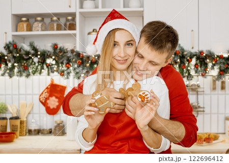 Loving husband hugs his sweet wife in santa hat from behind. Woman shows a gingerbread man in medical mask in one hand and tiger paw ginger cookie in another. Christmas and coronavirus concept Loving husband hugs his sweet wife in santa hat from behind. Woman shows a gingerbread man in medical mask in one hand and tiger paw ginger cookie in another. Christmas and coronavirus concept 122696412