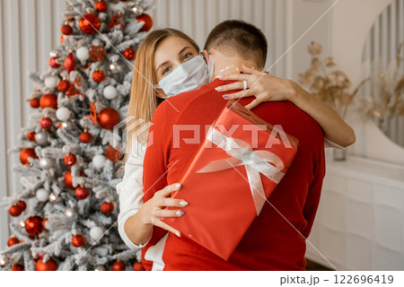 Close up of happy couple in medical masks with christmas gift hugging at home near Christmas tree 122696419