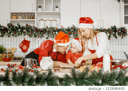 Happy cheerful family in santa hats cut out cookies from raw dough with a cookie cutter in the kitchen. Homemade cakes, Christmas sweets, the concept of winter holidays. Happy cheerful family in santa hats cut out cookies from raw dough with a cookie cutter in the kitchen. Homemade cakes, Christmas sweets, the concept of winter holidays. 122696493