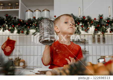 Happy fair-haired child in red sweater stands at the kitchen table with iron sieve in his hand at home looking proudly into camera. Happy fair-haired child in red sweater stands at the kitchen table with iron sieve in his hand at home looking proudly into camera. 122696513