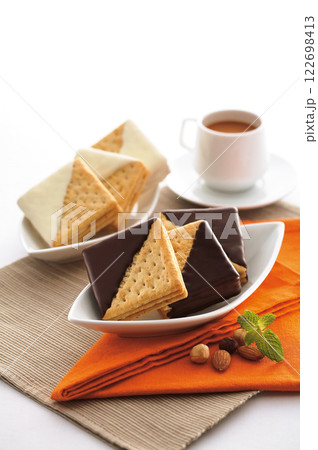 Cozy breakfast with a plate of crispy biscuits dipped in dark and white chocolate, served with a cup of steaming coffee. Garnished with nuts and mint leaves on a vibrant orange napkin. 122698413